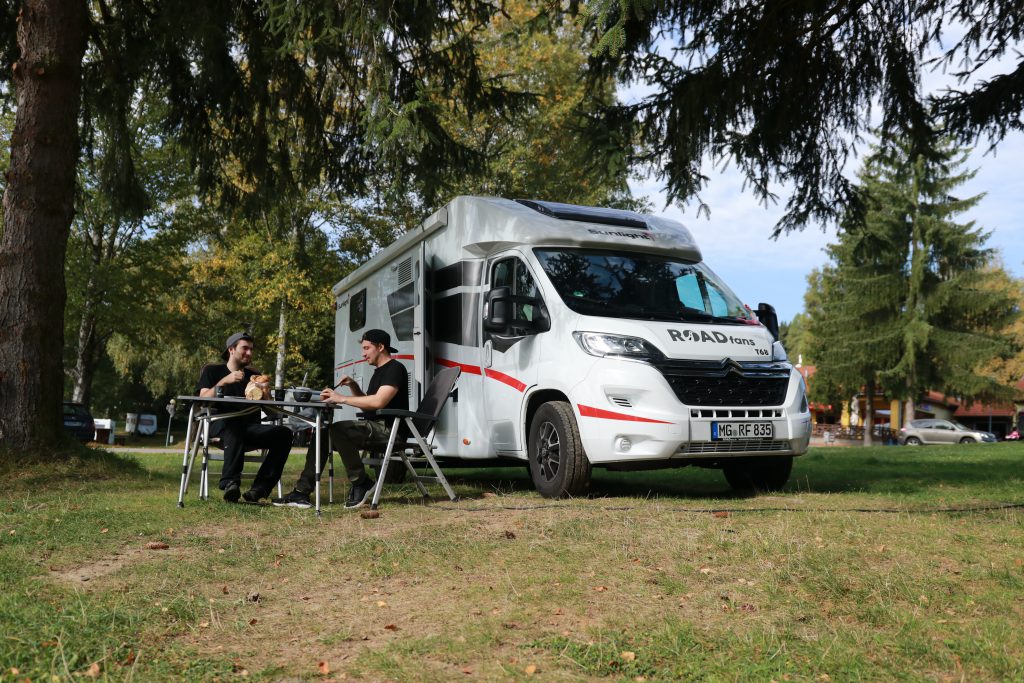 Zwei Freunde an Campingmöbeln vor dem Wohnmobil auf dem Campingplatz Köln