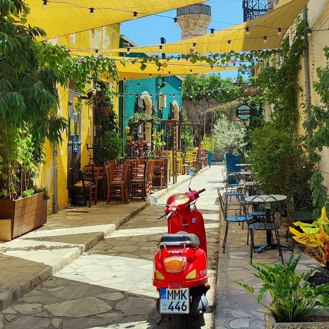 Roter Roller in einer Gasse mit kleinen Restaurants, links und rechts sind pflanzen und über der Gasse sind gelbe Laken gegen die Sonne gespannt.