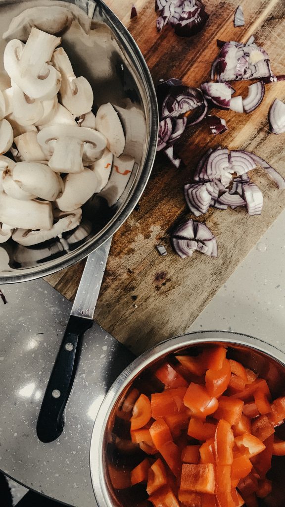 Nachhaltig Campen - Blick auf 2 Schüsseln, welche jeweils voll mit Champignons und mit Paprika sind. Im Hinter grund liegen auf einem Schneidebrett ein Messer und geschnittene Zwiebeln.