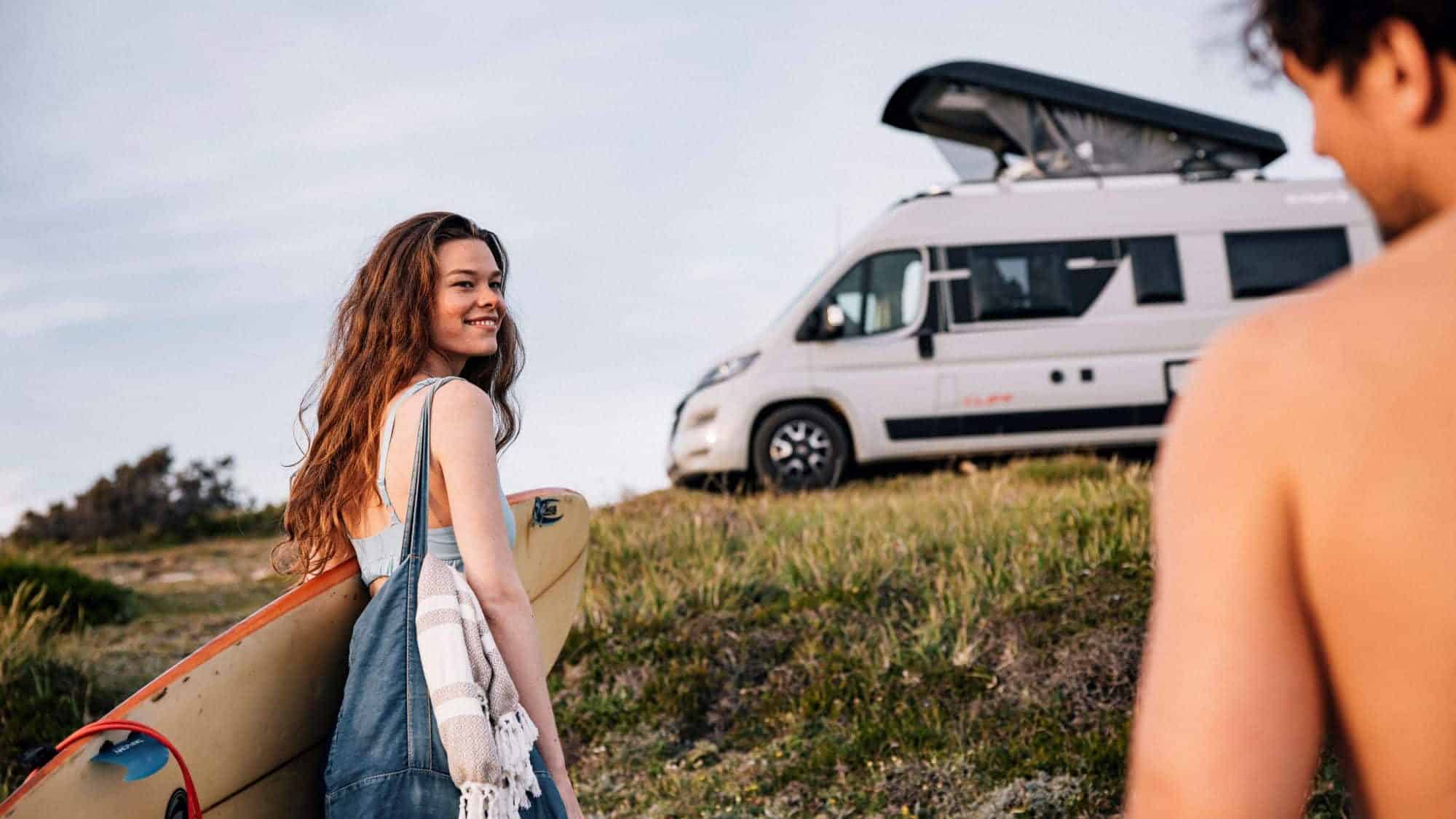 Frau mit Surfbrett lächelt Mann auf einer Wiese an. Im Hintergrund steht ein Camper mit Aufstelldach