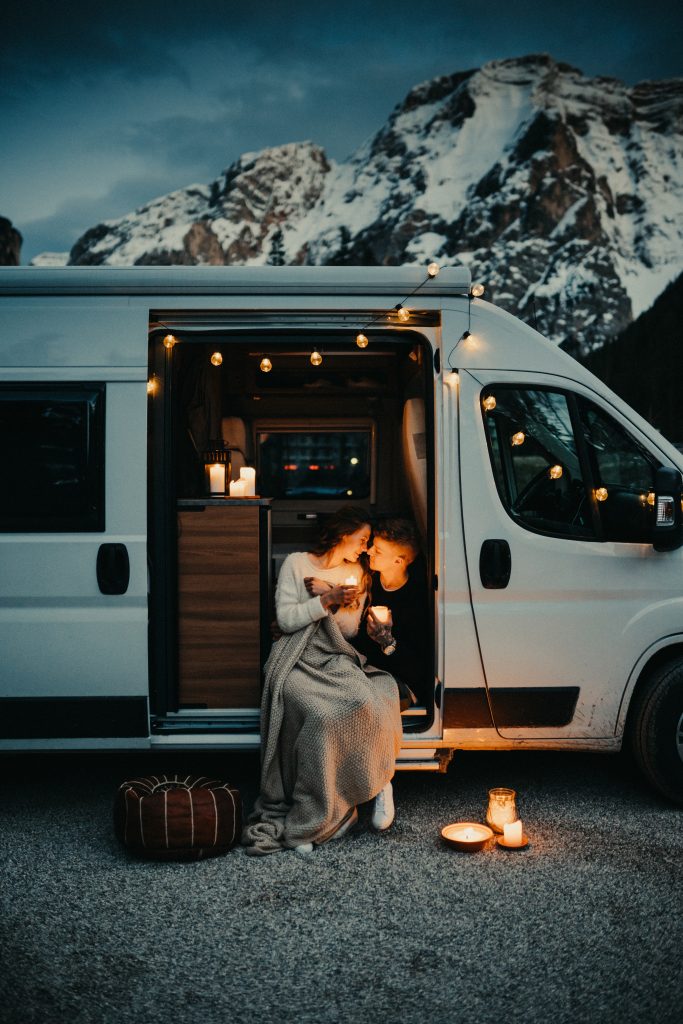 Weihnachten im Wohnmobil - Zwei Personen sitzen in der offenen Tür eines Wohnmobils, umgeben von Lichterketten, während sie gemeinsam eine Kerze halten, mit einem Bergpanorama im Hintergrund bei Dämmerung.