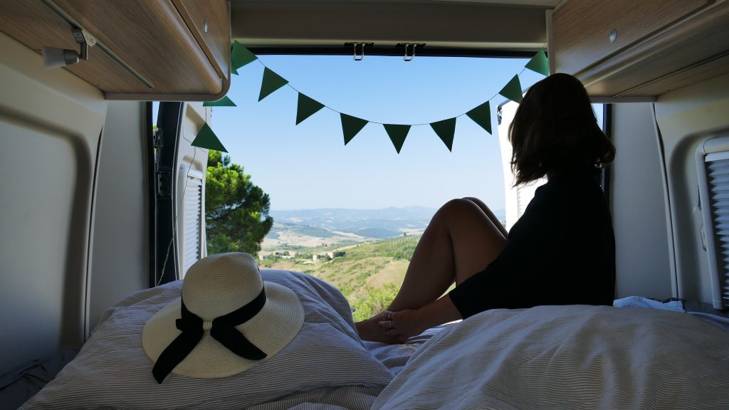 Wildcampen Strafen - Blick auf eine im Bett sitzende Frau, welche aus den geöffneten Wohnmobil Hecktüren in die Natur schaut.